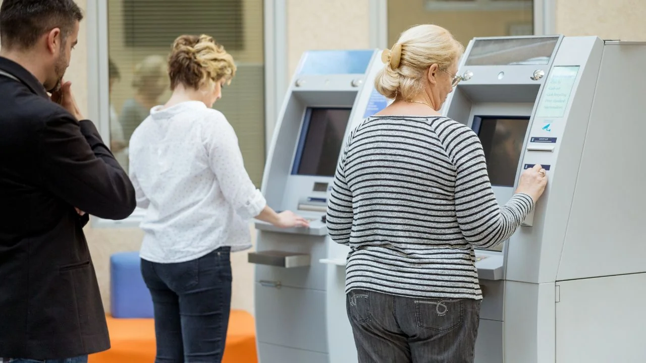 Wnętrze placówki bankowej z trzema osobami korzystającymi z samoobsługowych terminali i stojącymi obok urządzeń.